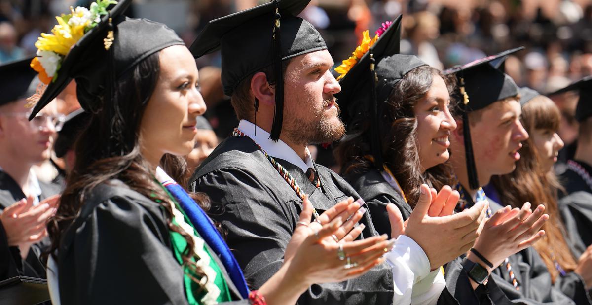 A row of graduates in black caps and gowns are standing and clapping, with rows of students and family members visible behind them.